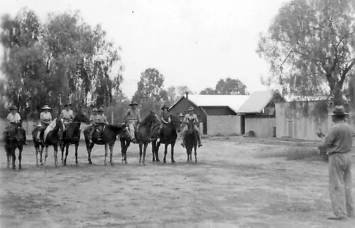 A black and white photograph of seven riders on horseback lined up in front of the station sheds